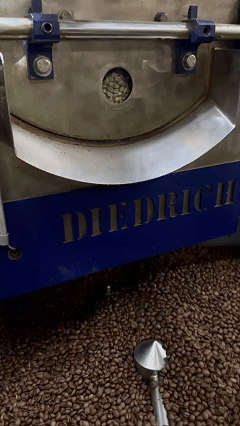 Тут видно два этапа: в смотровом окошке барабана — зеленое зерно внутри (еще не обжарено), а на охладительном лотке внизу — уже готовое темное зерно. 